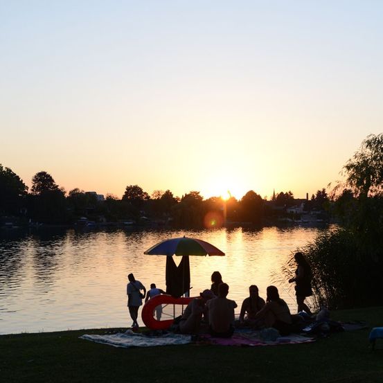 Eine Gruppe von Menschen genießt ein Picknick am See bei Sonnenuntergang, wobei eine Person einen Regenschirm hält.