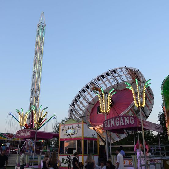 Ein farbenfroher Vergnügungspark mit einem hohen Riesenrad, einer Drehkarussell und verschiedenen Ständen. Menschen gehen herum, und der Himmel ist klar.
