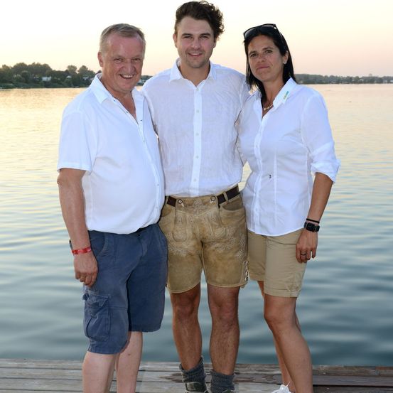 Drei Menschen stehen auf einem Steg am Wasser und lächeln. Der Mann links trägt ein rotes Armband, der Mann in der Mitte trägt Shorts und einen Gürtel und die Frau rechts trägt eine Uhr.