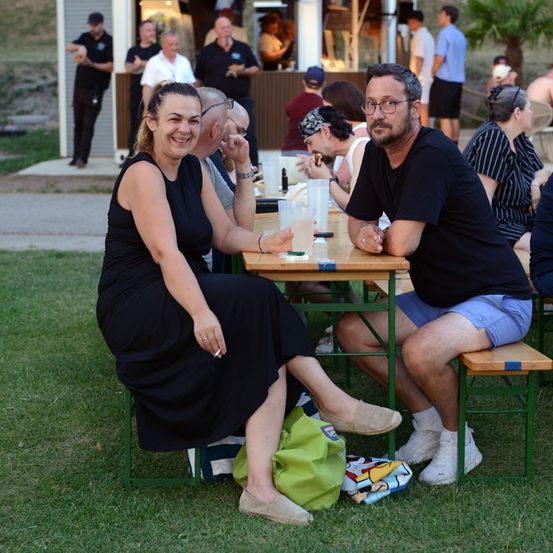 Eine Gruppe von Menschen sitzt an einem Tisch im Freien. Eine Frau lächelt, während ein Mann neben ihr etwas betrachtet. Andere Menschen sitzen an anderen Tischen, und einige stehen in der Nähe eines Imbissstandes.
