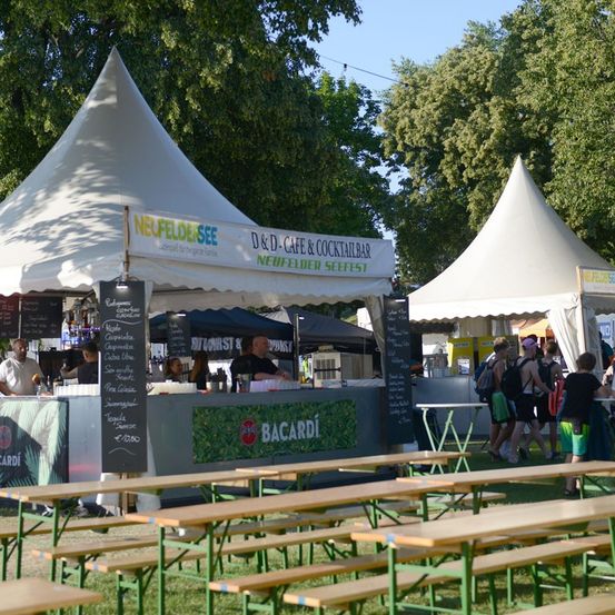 Ein Food-Festival mit weißen Zelten und Holzbänken, mit einem Menübrett und einem Bacardi-Schild. Leute gehen herum und einige stehen an der Theke.