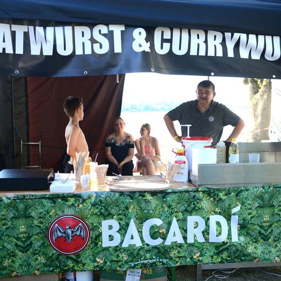 Ein Imbissstand mit einem Zeltdach, das 'WURST & CURRYWURST' bewirbt. Ein Mann steht hinter dem Tresen, während ein Junge und zwei Frauen in der Nähe sitzen. Flaschen und Behälter stehen auf dem Tresen, und ein Bacardi-Banner ist vorne.