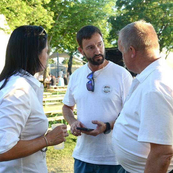 Drei Menschen in weißen Shirts stehen in einem Außenbereich, einer der Männer hält ein Telefon und schaut darauf. Die Frau auf der linken Seite hält einen Becher. Hinter ihnen sind Holzbänke und Bäume.