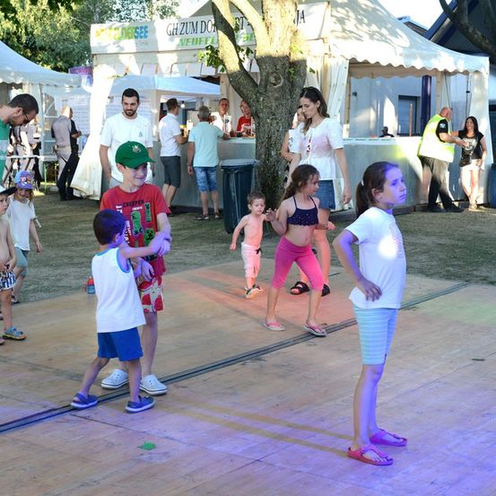 Eine Gruppe von Kindern und Erwachsenen tanzt auf einer hölzernen Bühne mit bunten Lichtern bei einer Veranstaltung im Freien.