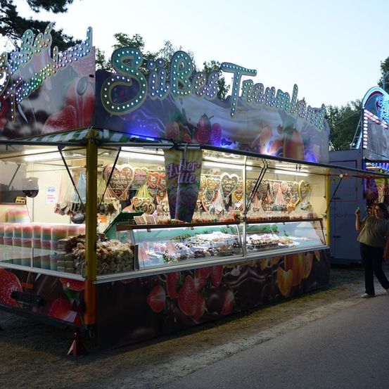 Ein leuchtender Imbissstand mit einem Neonschild, das 'Silke Traumwelten' anzeigt, präsentiert eine Auswahl an Desserts, darunter herzförmige Kekse und schokoladeüberzogene Erdbeeren. Eine Person geht vorbei.
