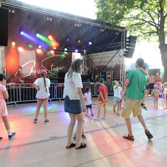 Eine Gruppe von Kindern und Erwachsenen tanzt auf einer Bühne mit bunten Lichtern und einem Baum im Hintergrund.