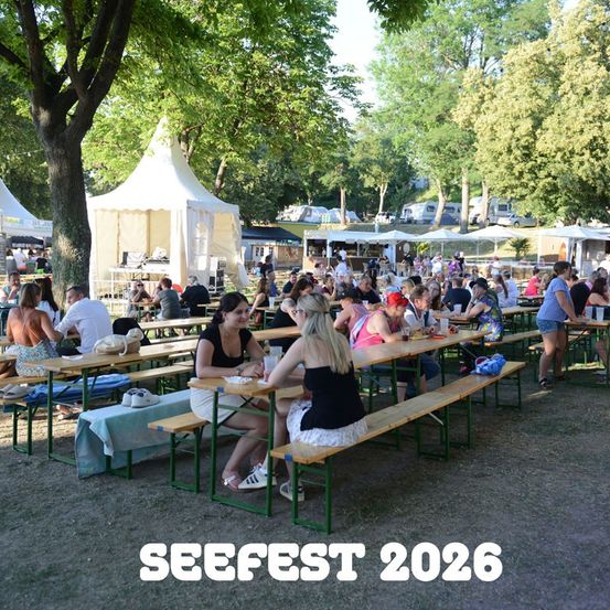 Ein geschäftiges Outdoor-Festival mit Menschen, die an Tischen unter Zelten sitzen und Speisen und Getränke genießen. Bäume und geparkte Fahrzeuge sind im Hintergrund zu sehen. Das Bild ist mit 'SEEFEST 2026' beschriftet.