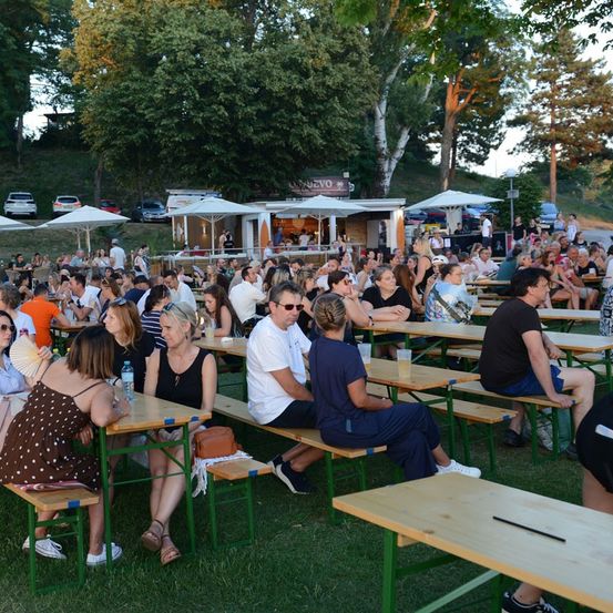 Eine große Menschenmenge sitzt an Tischen im Freien, möglicherweise bei einem Festival. Einige tragen Sonnenbrillen und Hüte. Im Hintergrund befinden sich Zelte und Bäume.