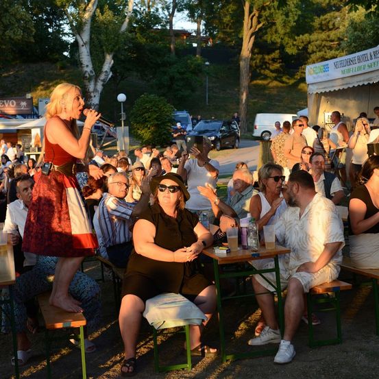 Eine Frau in einem roten Kleid singt in ein Mikrofon bei einer Outdoor-Veranstaltung, während Menschen an Tischen und Bänken sitzen und zuhören.