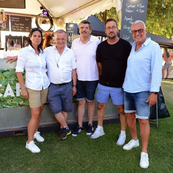 Fünf Personen stehen lächelnd auf einer Wiese für ein Foto. Sie sind lässig gekleidet, zwei Männer tragen Brillen und einer trägt eine Uhr. Hinter ihnen ist ein Zelt mit einem Schwarzen Brett zu sehen, auf dem Getränkepreise stehen.