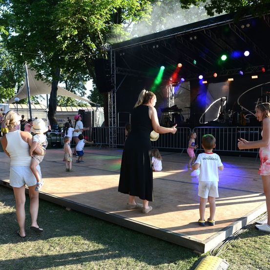 Eine Gruppe von Erwachsenen und Kindern versammelt sich um eine Bühne mit bunten Lichtern. Eine Frau in einem schwarzen Kleid steht auf der Bühne, während andere zuschauen und interagieren.