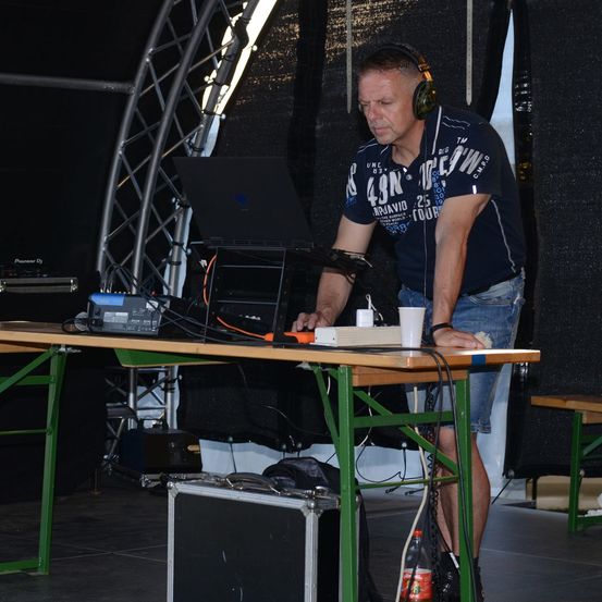 Ein Mann mit Kopfhörern und einem blauen T-Shirt steht an einem Tisch mit Laptop und anderem Equipment. Er bereitet sich wahrscheinlich auf eine Aufführung vor.