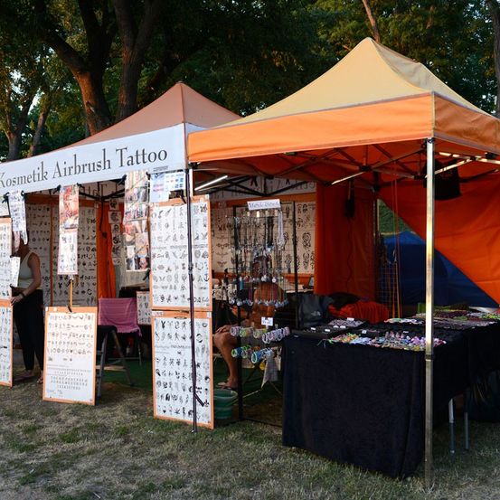 Zwei Zelte mit orangefarbenen Zeltdächern auf einem Rasen aufgestellt, eines mit der Aufschrift 'Kosmetik Airbrush Tattoo' mit verschiedenen Designs zur Schau gestellt und ein anderes mit einem Tisch voller Schmuck. Eine Person sitzt unter einem der Zelte.