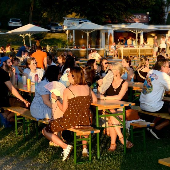 Eine Gruppe von Menschen sitzt an Tischen im Freien unter Schirmen, einige halten Fächer, in einer lebhaften Umgebung im Freien.