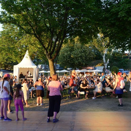 Eine Gruppe von Menschen versammelt sich in einem Außenbereich, wahrscheinlich für ein Festival. Einige sitzen auf Stühlen um Tische, während andere stehen. Eine Frau in einem roten Kleid steht auf der Bühne, möglicherweise bei einer Aufführung. Es gibt Zelte und ein weißes Vordach im Bereich.