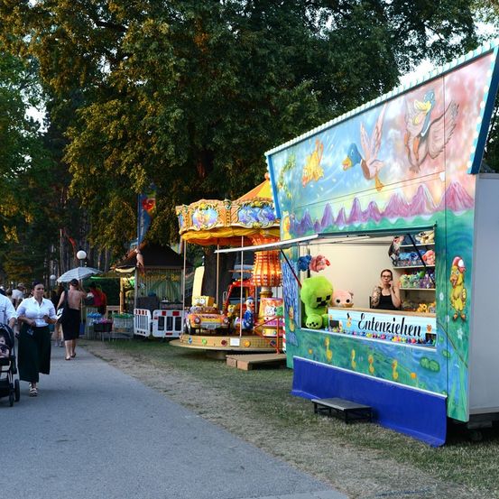 Eine Straßenszene mit einem farbenfrohen Jahrmarktstand, der Spielzeug verkauft. Menschen gehen auf einem Bürgersteig vorbei, und eine Frau telefoniert im Stand.