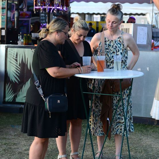 Drei Frauen stehen um einen Tisch bei einer Outdoor-Veranstaltung, eine hält ein Getränk, eine andere schaut nach unten und die dritte hält eine Handtasche.