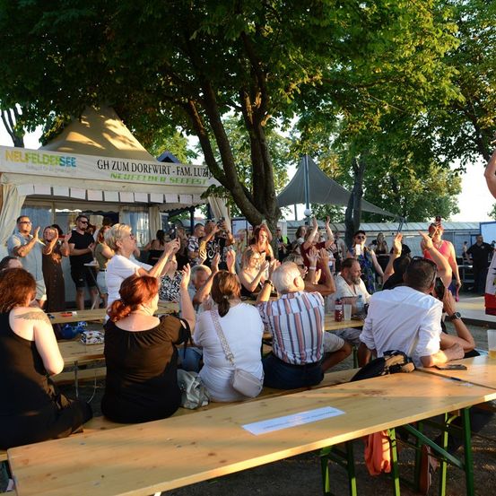 Eine Gruppe von Menschen versammelt sich im Freien unter einem Baum, klatscht und jubelt, möglicherweise bei einem Festival.