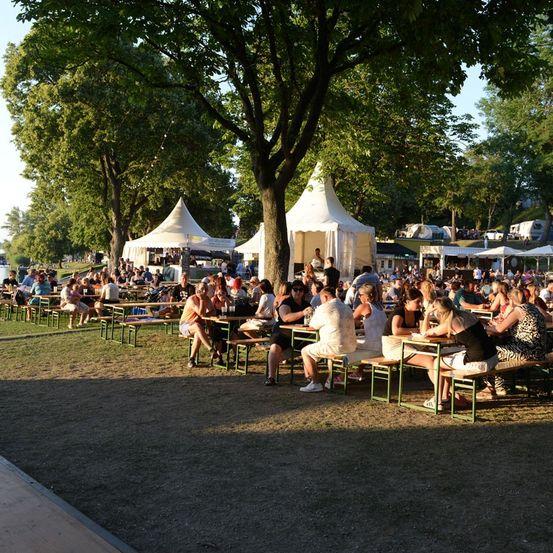 Eine große Gruppe von Menschen versammelt sich in einem Außenbereich, wahrscheinlich einem Park, für eine Veranstaltung. Sie sitzen an Tischen unter Zelten und Bäumen, einige Leute stehen herum.