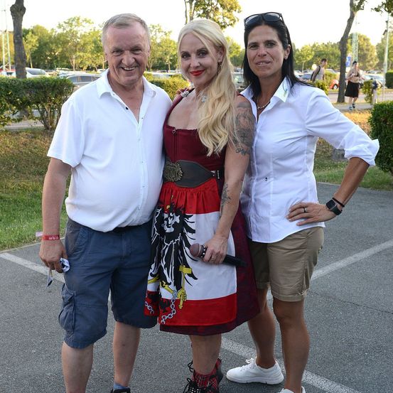 Drei Menschen stehen zusammen auf einem Parkplatz. Ein Mann in einem weißen Hemd und blauen Shorts, eine Frau in einem rot-weißen Kleid und eine Frau in einem weißen Hemd und hellbraunen Shorts.