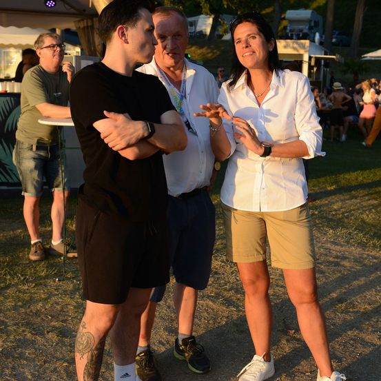 Drei Menschen stehen in einer Außenumgebung, wobei ein Mann seine Arme verschränkt und ein anderer Mann mit einer Frau in einer weißen Bluse und khakifarbenen Shorts spricht.