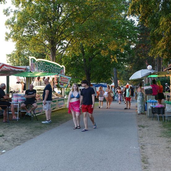 Eine Gruppe von Menschen geht auf einem Weg in einem Park mit Essensständen und Bäumen. Ein Paar im Vordergrund trägt Bikinis und Sandalen.