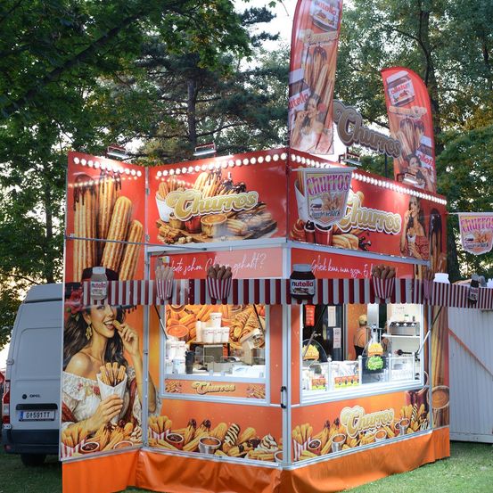 Ein Churro-Imbisswagen mit einer Frau an der Seite, der Churros und andere Desserts anbietet. Der Wagen ist mit bunten Grafiken dekoriert und hat ein rot-weiß gestreiftes Vordach.