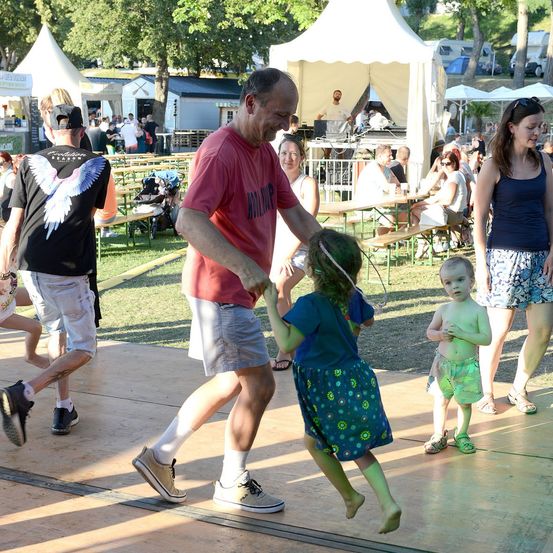 Ein Mann tanzt mit einem kleinen Mädchen auf einer Bühne bei einer Outdoor-Veranstaltung, während ein Kleinkind und eine Frau in der Nähe stehen. Im Hintergrund sitzen Leute an Tischen unter Zelten.