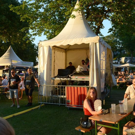 Eine Gruppe von Menschen genießt eine Musikveranstaltung in einem Park. Eine Frau sitzt an einem Tisch, während andere um einen DJ-Stand herumstehen. Bäume und Zelte sind im Hintergrund.