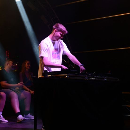 Ein Mann steht an einer DJ-Konsole, während eine Menge zuschaut, beleuchtet von einem Scheinwerfer.