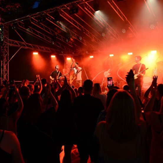 Eine Band spielt auf der Bühne mit roter und gelber Beleuchtung, vor ihnen steht eine Menge von Menschen.