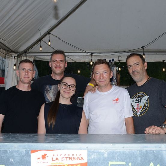 Fünf Personen stehen unter einem Zelt und lächeln für ein Foto. Eine Frau in der Mitte trägt eine Brille. Dahinter steht ein Schild mit der Aufschrift 'Pizzeria La Strega'.