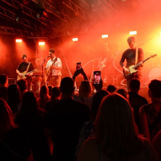 Eine Band spielt auf einer Bühne unter roten und gelben Lichtern, vor einer Menge von Menschen. Die Bandmitglieder spielen Gitarre und singen.