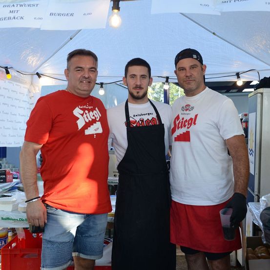 Drei Männer stehen unter einem Zelt, zwei tragen rote T-Shirts mit dem Wort 'Stiegl' und einer trägt eine schwarze Schürze. Der Mann in der Mitte hält einen Becher.