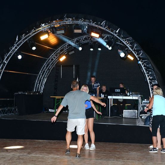 Eine nächtliche Veranstaltung mit einer Bühne, auf der eine Frau und ein Mann tanzen, während andere zuschauen. Die Bühne ist mit Scheinwerfern ausgestattet und ein DJ ist anwesend.