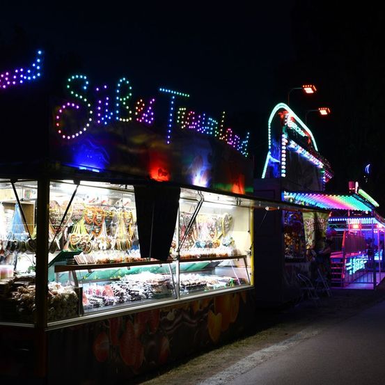 Ein hell beleuchteter Jahrmarktstand bei Nacht mit bunten Lichtern und einem Schild, das 'Salz & Geschmacksland' besagt. Der Stand ist mit verschiedenen Süßigkeiten und Leckereien gefüllt.