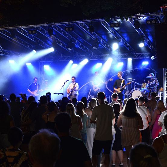 Eine Band spielt auf der Bühne unter blauen und weißen Lichtern vor einer Menge bei Nacht.