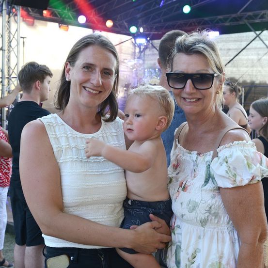 Zwei Frauen und ein Kleinkind stehen bei einer Outdoor-Veranstaltung zusammen und lächeln für die Kamera. Das Kleinkind hält sich an der Frau auf der linken Seite fest, während die Frau auf der rechten Seite eine Sonnenbrille trägt. Hinter ihnen stehen Menschen und beobachten eine Bühne mit bunten Lichtern.
