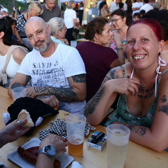 Eine Gruppe von Menschen sitzt an einem Tisch in einer Außenumgebung. Ein Mann und eine Frau lächeln und schauen zur Kamera. Der Mann trägt ein weißes T-Shirt mit einem grafischen Design. Die Frau trägt ein ärmelloses Oberteil und hat Tattoos an den Armen. Auf dem Tisch stehen Gläser, eine Zigarettenpackung und ein Teller mit Essen.