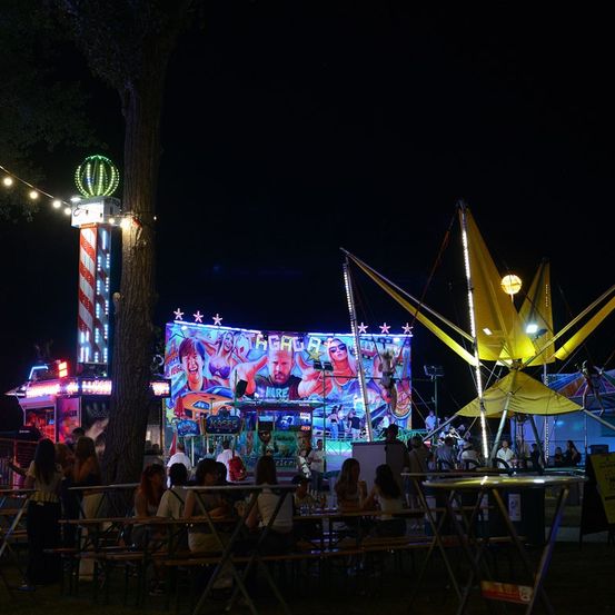 Eine Nachtszene auf einem Jahrmarkt mit Menschen, die an Tischen unter einem großen beleuchteten Baldachin sitzen und verschiedene Fahrgeschäfte im Hintergrund.