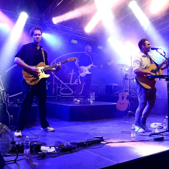 Drei Musiker spielen auf einer Bühne mit lila Beleuchtung, Gitarren und einem Mikrofon. Der Mann links spielt eine braune Gitarre, der in der Mitte eine weiße Gitarre und der rechts eine braune Gitarre.