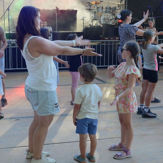 Eine Gruppe von Kindern und Erwachsenen tanzt auf einer Bühne mit einer Band im Hintergrund. Die Frau vorne führt den Tanz an.