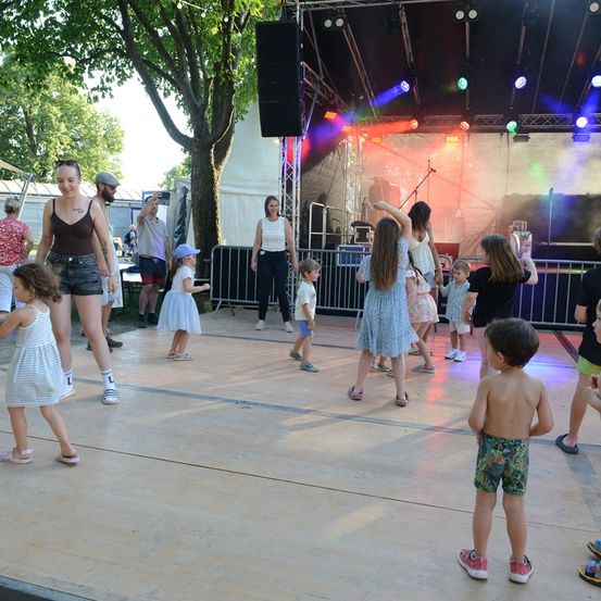 Eine Gruppe von Menschen, einschließlich Kindern, tanzt auf einer Bühne mit bunten Lichtern und einem Baum im Hintergrund.
