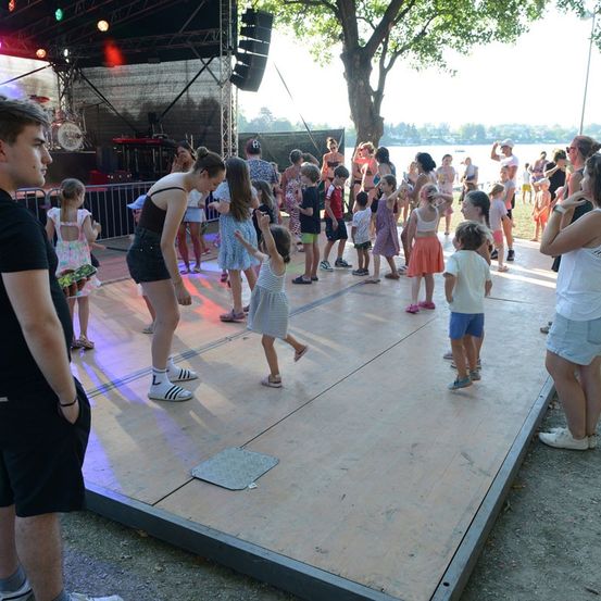 Eine Gruppe von Kindern und Erwachsenen tanzt auf einer Bühne bei einer Outdoor-Veranstaltung, mit einer malerischen Kulisse eines Sees und Bäumen.