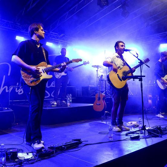 Vier Musiker spielen auf der Bühne mit Gitarren unter blauer Bühnenbeleuchtung. Ein Mann singt in ein Mikrofon, während ein anderer Gitarre spielt.