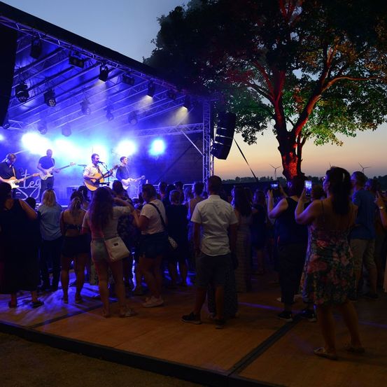 Eine Band spielt auf einer Freiluftbühne bei Dämmerung, während eine Menge Menschen zusieht und einige Fotos machen. Die Bühne ist mit blauen und lila Lichtern beleuchtet, und in der Nähe steht ein Baum mit roten und grünen Lichtern.