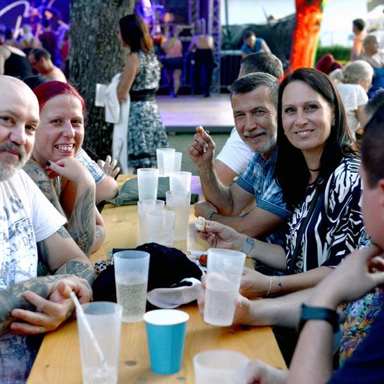 Eine Gruppe von Menschen sitzt an einem Tisch mit Gläsern und Essen, lächelt und genießt eine Veranstaltung im Freien.