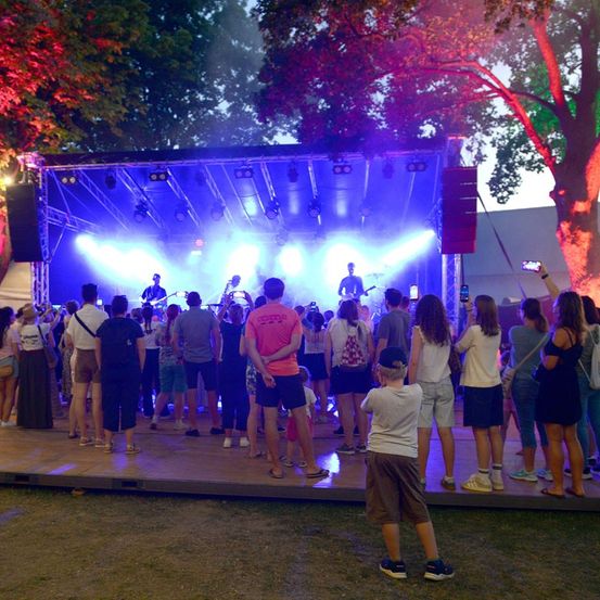 Ein nächtliches Open-Air-Konzert mit einer Band auf der Bühne, beleuchtet von blauen und roten Lichtern, und einer Menge von Zuschauern.