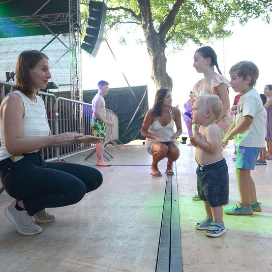 Eine Frau hockt auf einer Bühne mit einer Gruppe von Kindern, von denen eines eine Puppe hält. Dahinter hockt eine Frau und interagiert mit einem anderen Kind. Im Hintergrund steht ein Junge in der Nähe eines Baumes und eines Metallzauns.