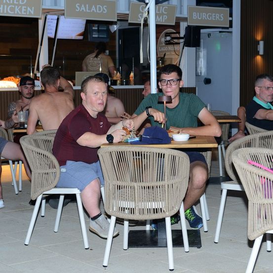 Zwei Männer sitzen an einem Tisch in einem ungezwungenen Essbereich, einer trägt eine Brille und der andere eine Uhr. Hinter ihnen weisen Schilder auf Speiseoptionen wie Hähnchen, Salate und Burger hin.
